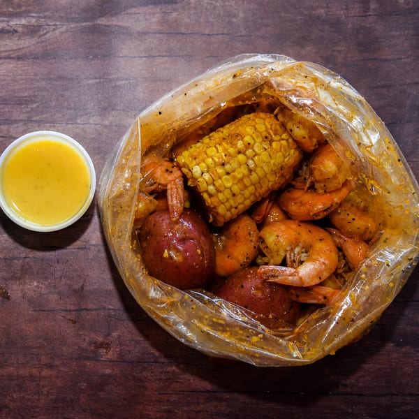 Shrimp, corn, and potatoes seasoned in a clear bag with a side of yellow dipping sauce.